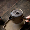 A hand grips the handle of a matte black teapot with a beige rim on a wooden coaster.