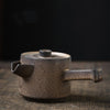 Side profile of a matte black teapot with a beige rim on a wooden surface, dark tone.
