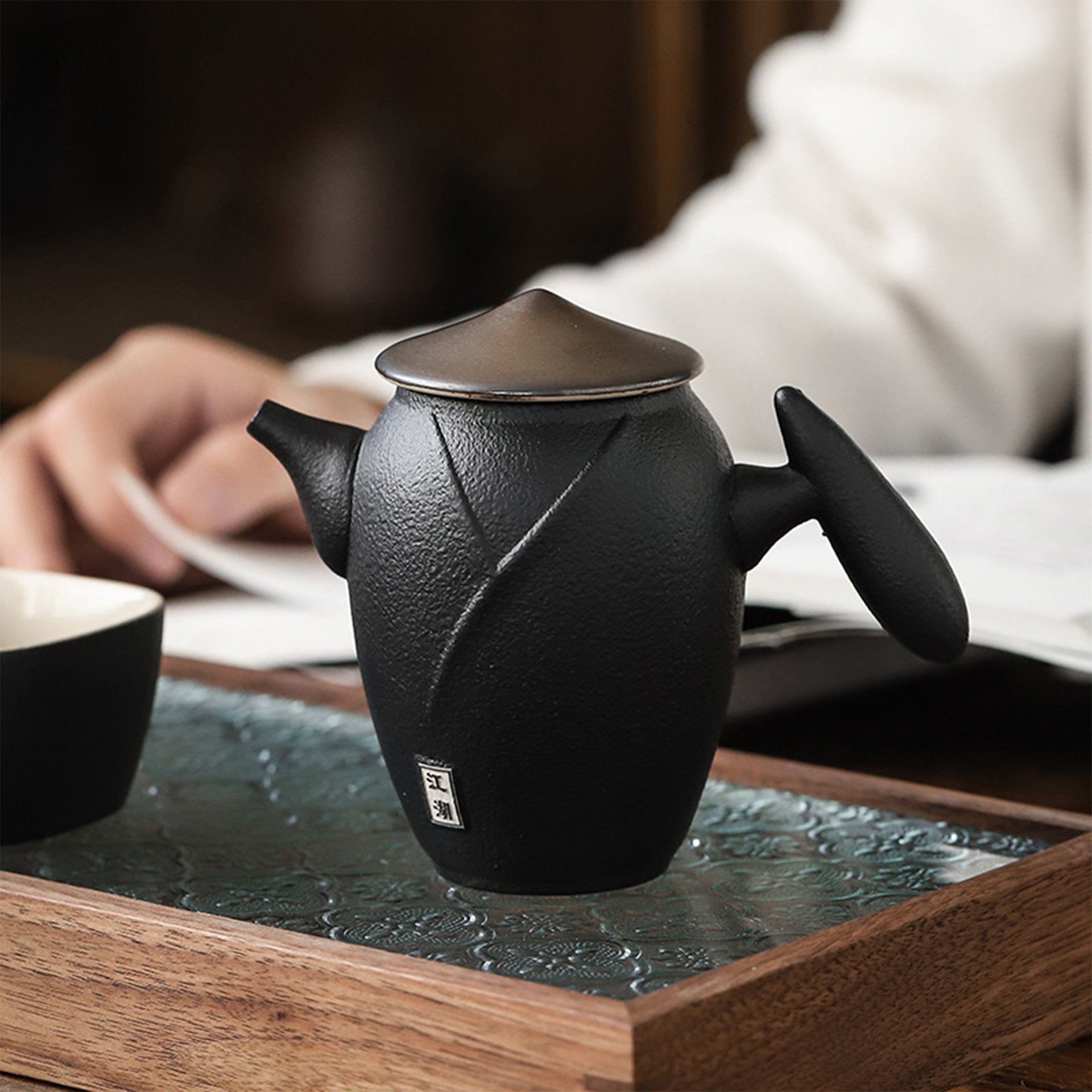 Matte black teapot with lid on wooden tray, person reading in the background.