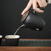 Hand tilting black teapot pouring tea into cup, dark backdrop.