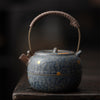 Full view of a rustic, blue and bronze-glazed teapot on a wooden surface in dim light.