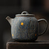 Dark blue clay teapot with intricate texture and mustard yellow spots, standing on a wooden ledge.