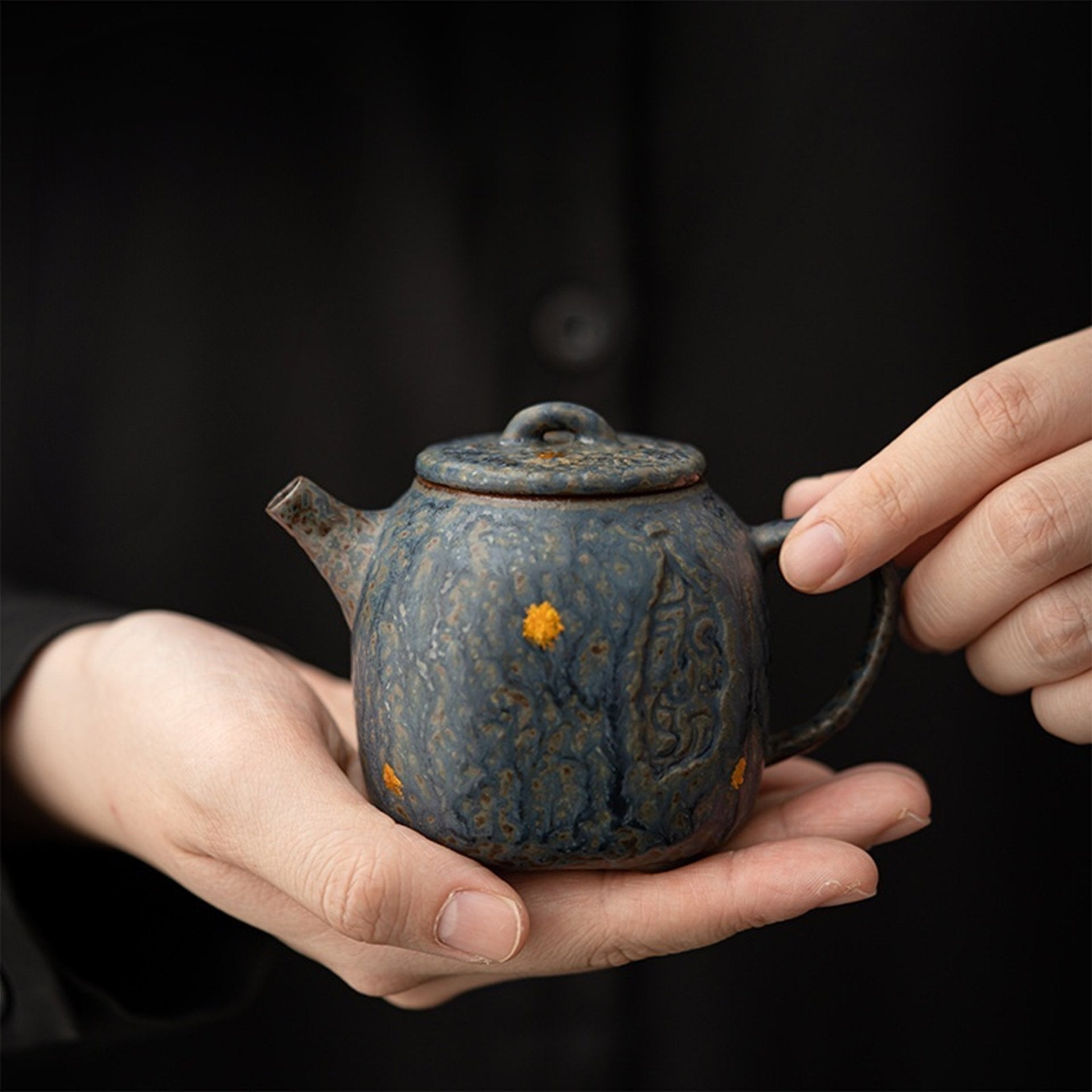 Two hands cradling a small textured blue clay teapot with yellow speckles on a black backdrop.