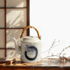 White teapot with a black swirl, placed on a bamboo mat next to dry branches.