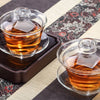 Two glass teapots with amber tea on a patterned mat.