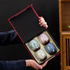Open gift box held by hand, showcasing four glazed teacups with pastel and earthy tones against a dark backdrop.