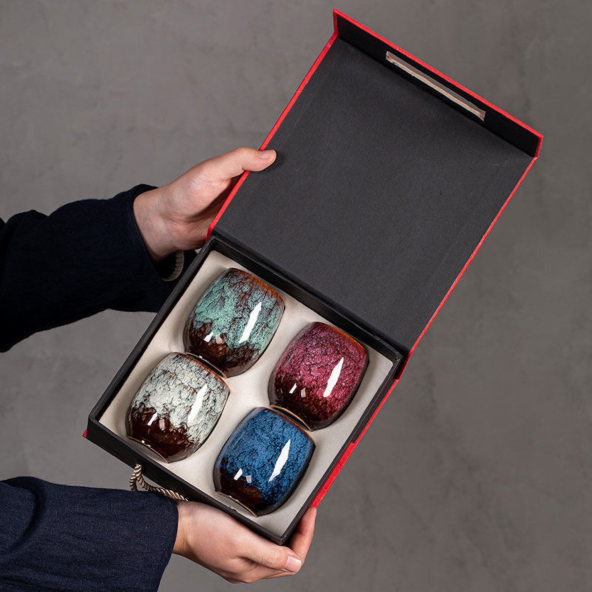Hand holding open box containing four unique glazed teacups in rich, earthy tones.