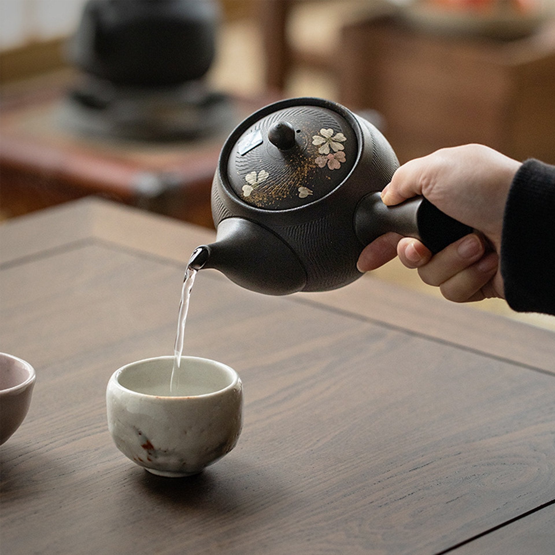 Hand pouring water from a traditional dark teapot into a cup.