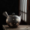 Dark teapot with floral design on a wooden surface, against a dark background.