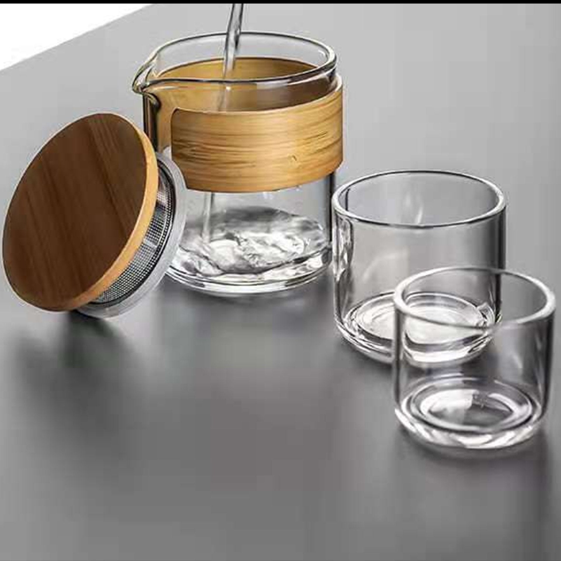 Glass tea infuser with bamboo accents being filled, next to empty glasses.