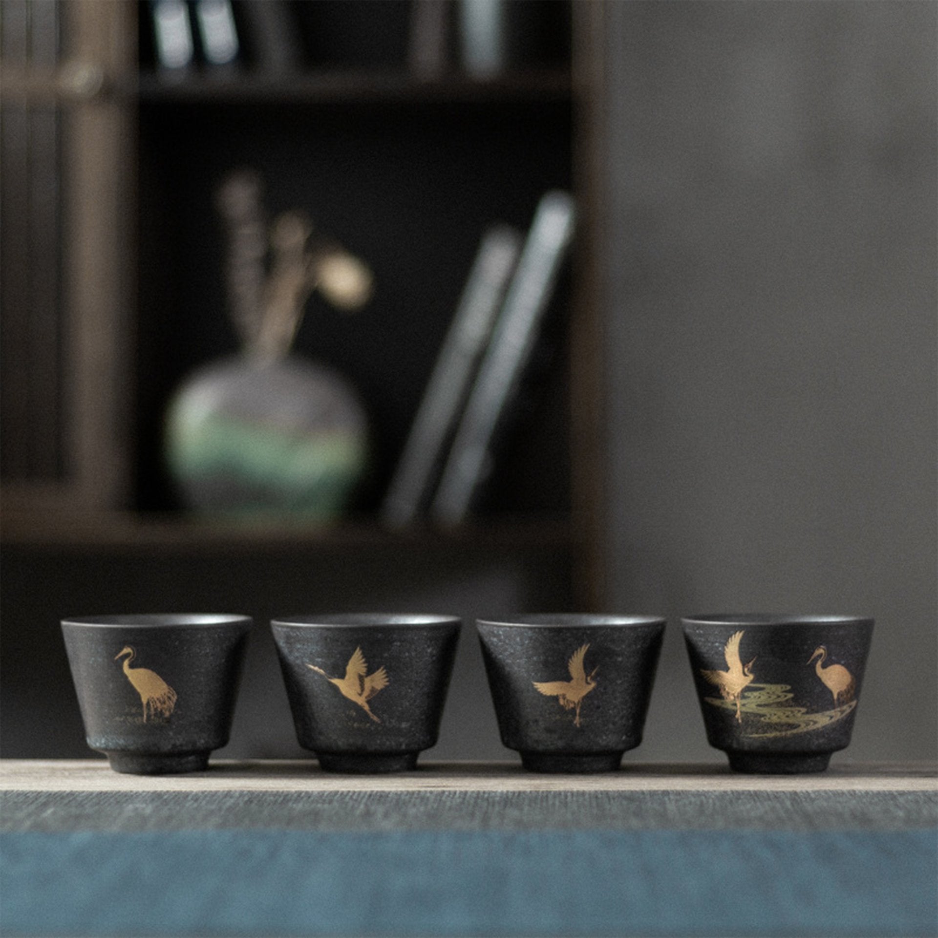 Four dark teacups with golden crane designs displayed on a blue cloth with a blurred bookshelf in the background