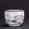 A white Japanese tea cup with traditional landscape artwork, featuring mountains, waves, a house, and a boat, with gold trim along the rim against a dark background.