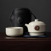A minimalist Japanese tea set featuring a cream teapot with a wooden lid knob, matching cup, navy cloth, and a sleek black carrying case.