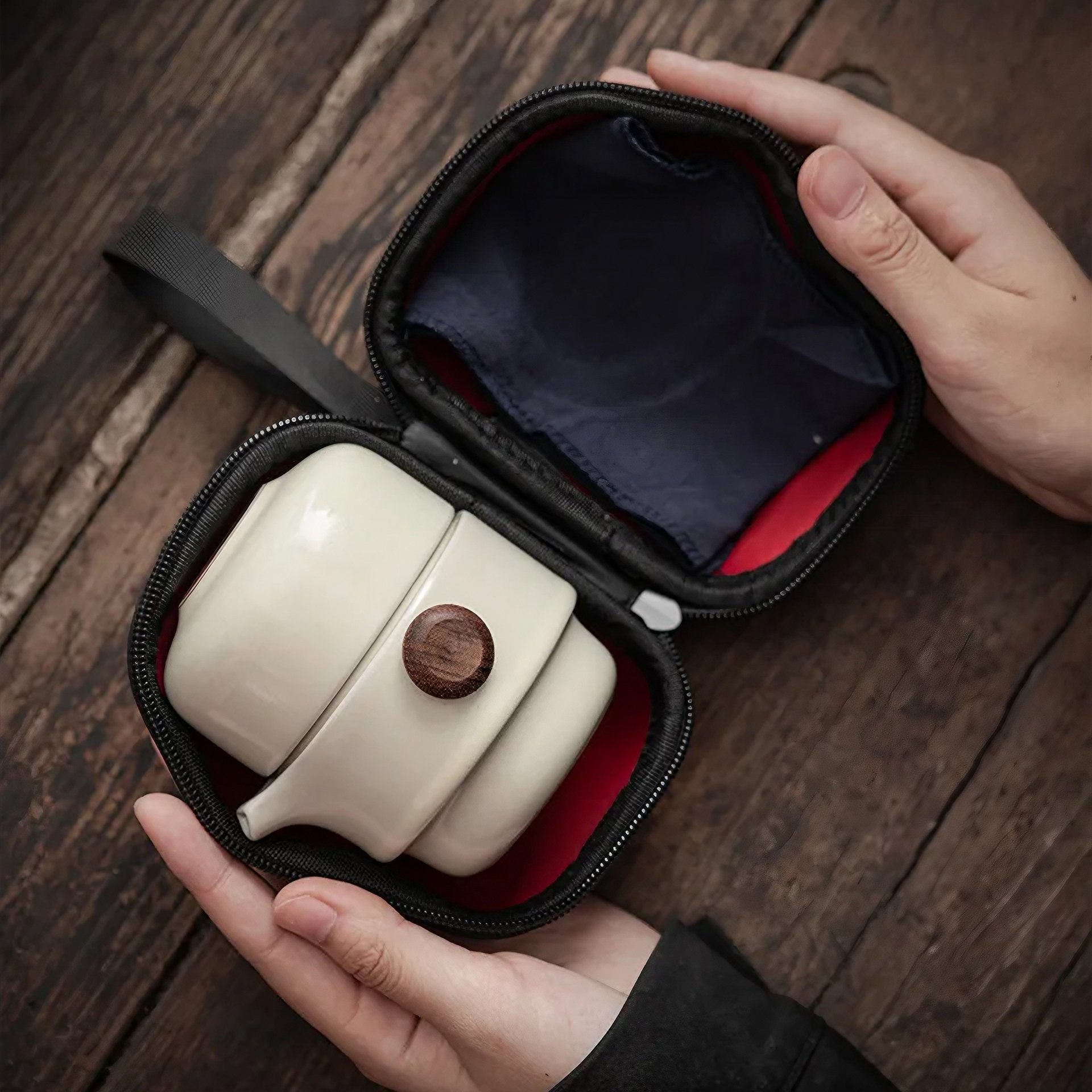 A hand holding a black zippered case open to reveal a minimalist white travel tea set, including a teapot with a circular dark wooden lid and two small cups, against a wooden surface.