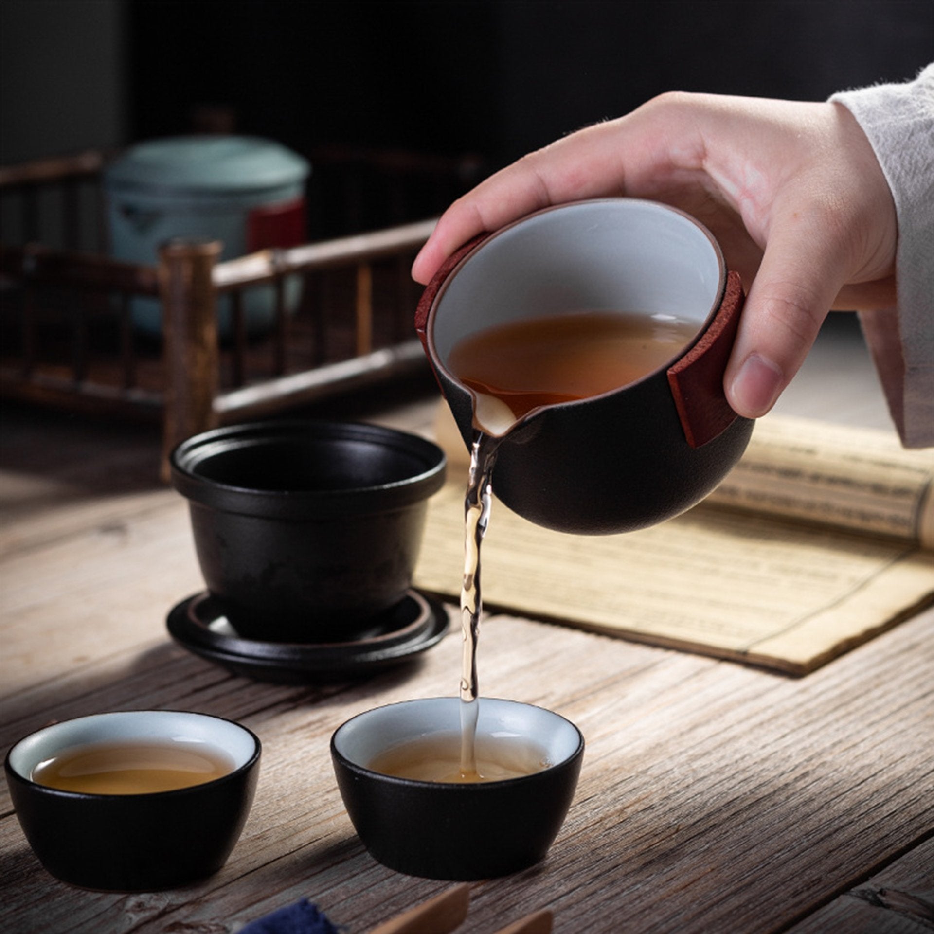 Hand pouring tea from a black pot into a cup on a wooden table with more tea accessories in the background.
