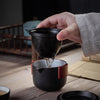 Hand pouring water from a black cup into a matching teapot on a wooden table with traditional tea ceremony items nearby.
