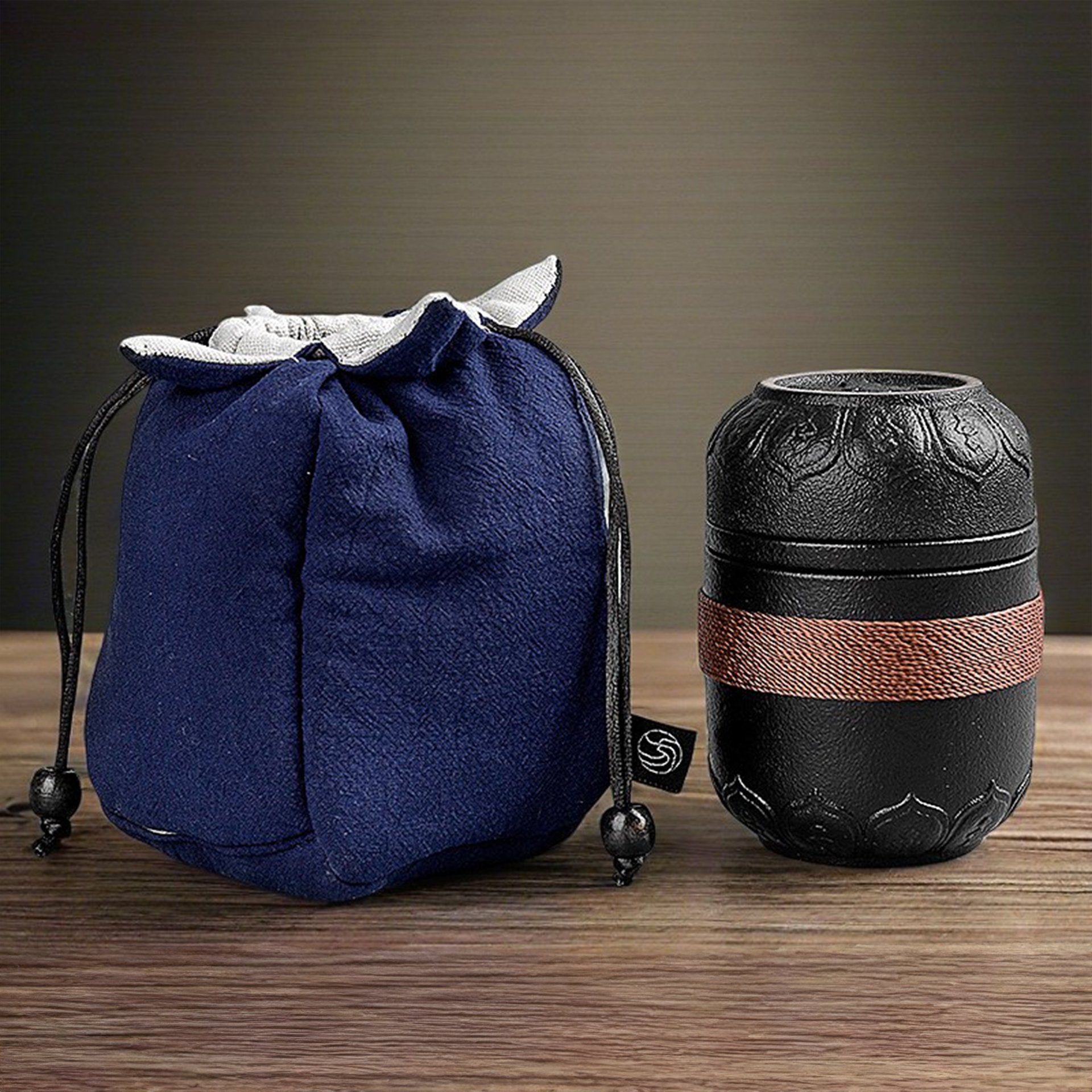 A black portable teapot with a copper band next to a blue fabric drawstring bag on a wooden surface.