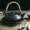 A dark, matte teapot with a bamboo handle and a wooden spoon of tea leaves on a table.