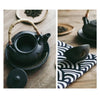 A black teapot with a bamboo handle on a plate, next to a cloth with a wave pattern.