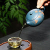Hand pouring tea from a blue floral-patterned teapot into a glass cup.