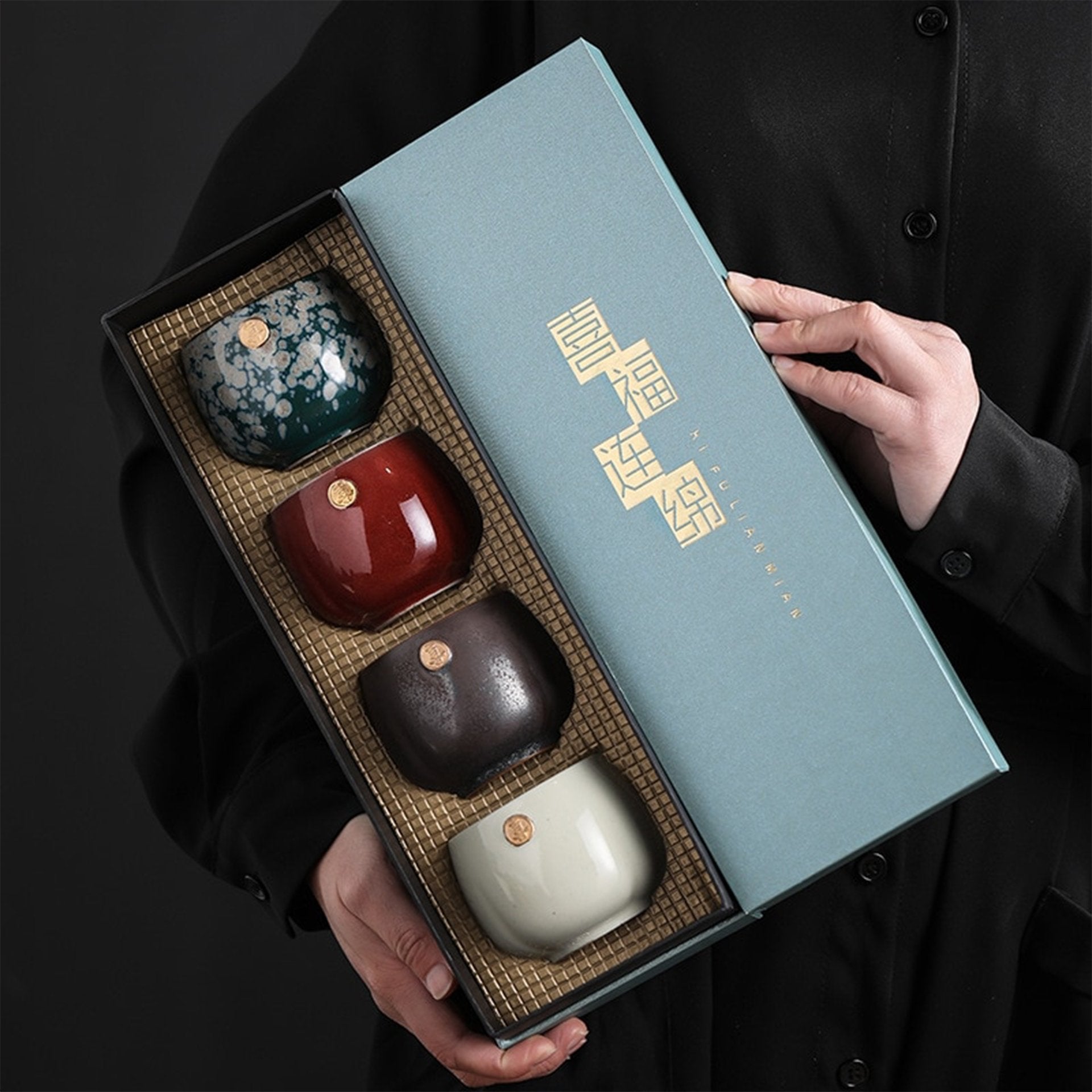 Person holding a gift box containing four Japanese-style teacups with gold seals.
