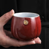 Hand holding a crimson red Japanese teacup with a gold seal.