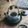 A hand scooping tea leaves into a teapot with a wave design and bamboo handle.