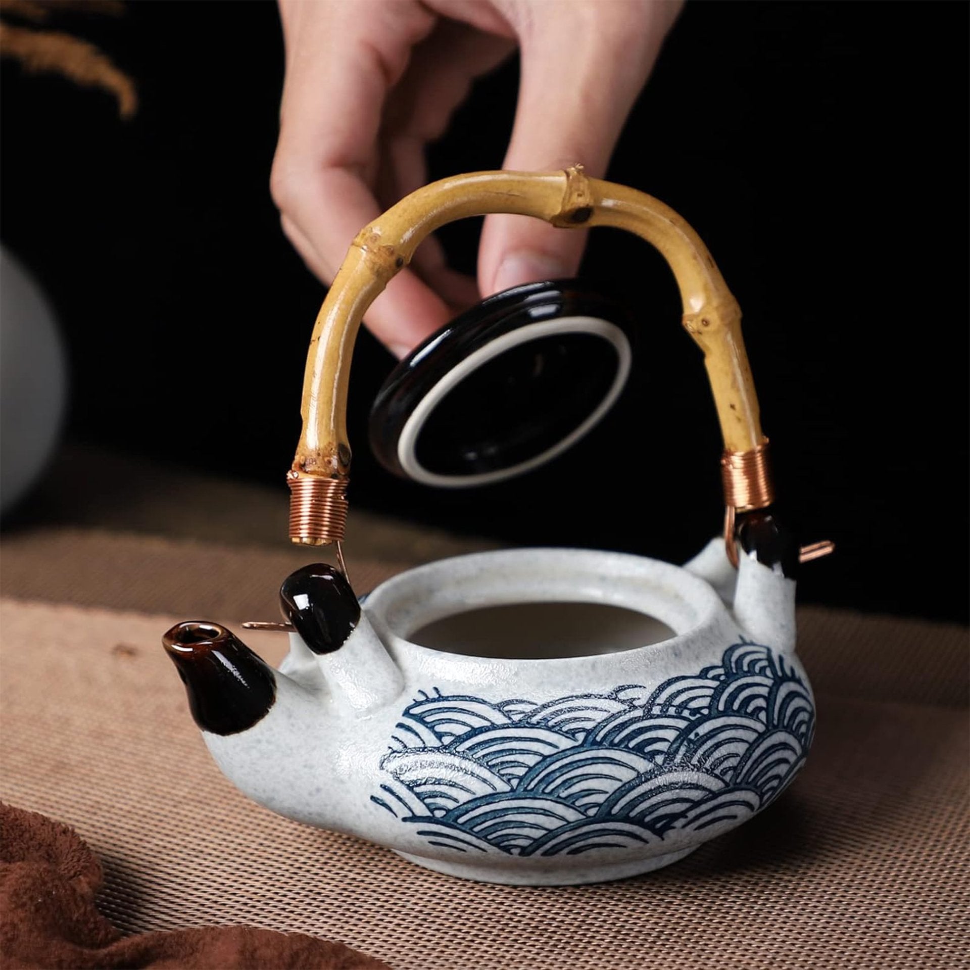 A hand lifting the lid off a patterned teapot with a bamboo handle.