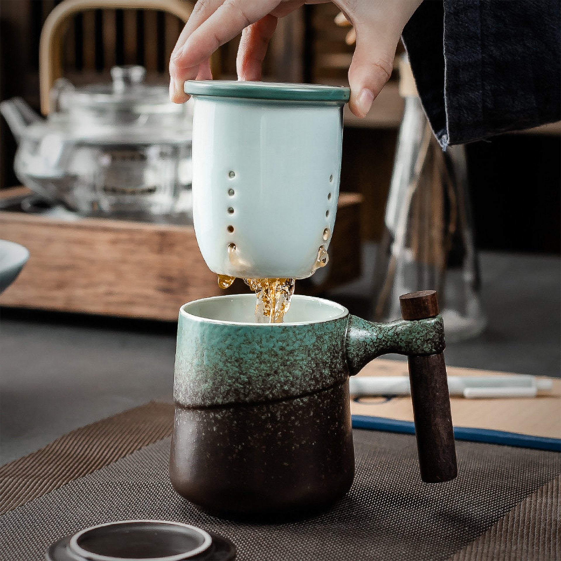 Tea being poured from a ceramic infuser into a matching dark green mug on a textured mat.