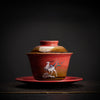 Side view of a red gaiwan on a saucer with deer imagery.