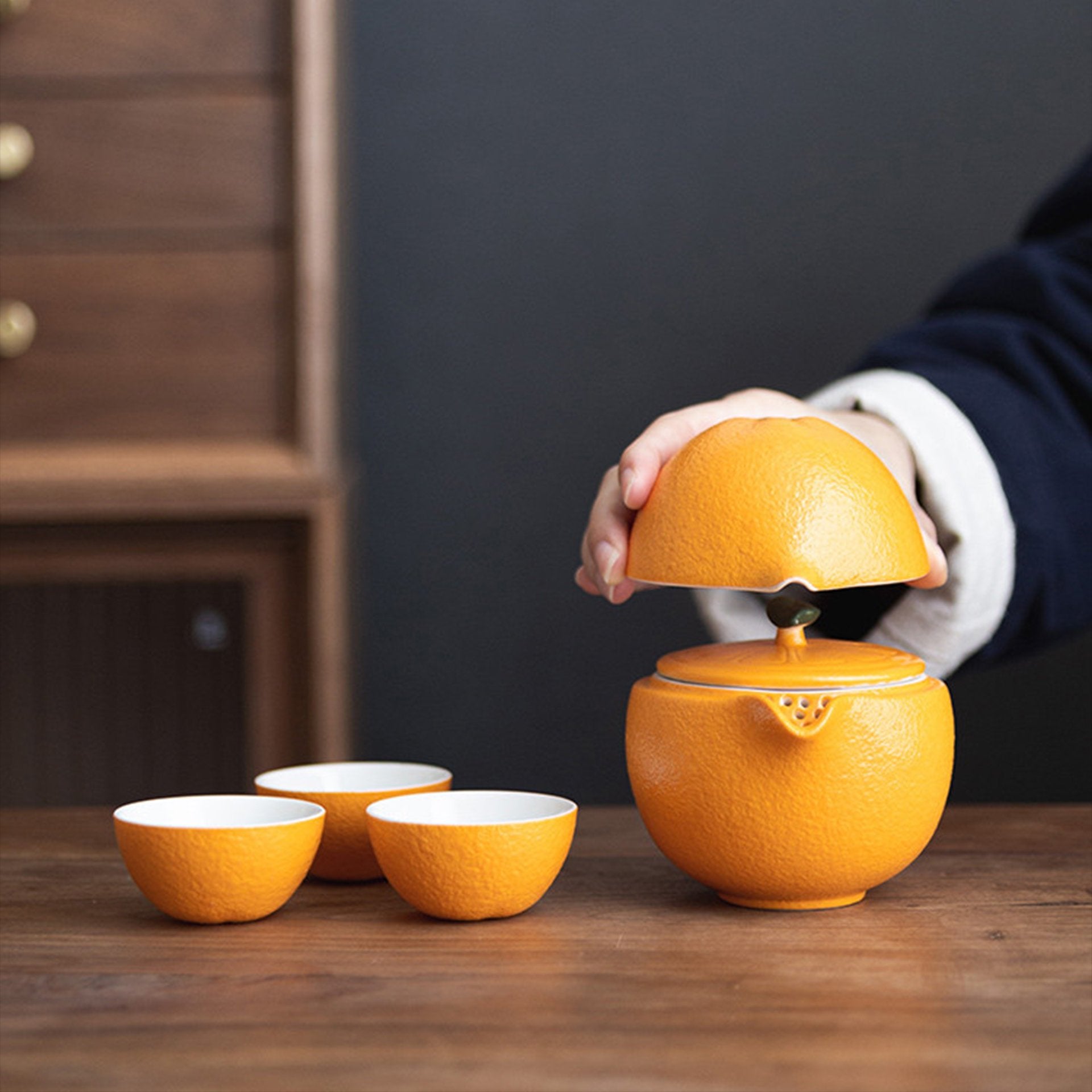 Hand lifting the lid of an orange ceramic teapot with tea cups.