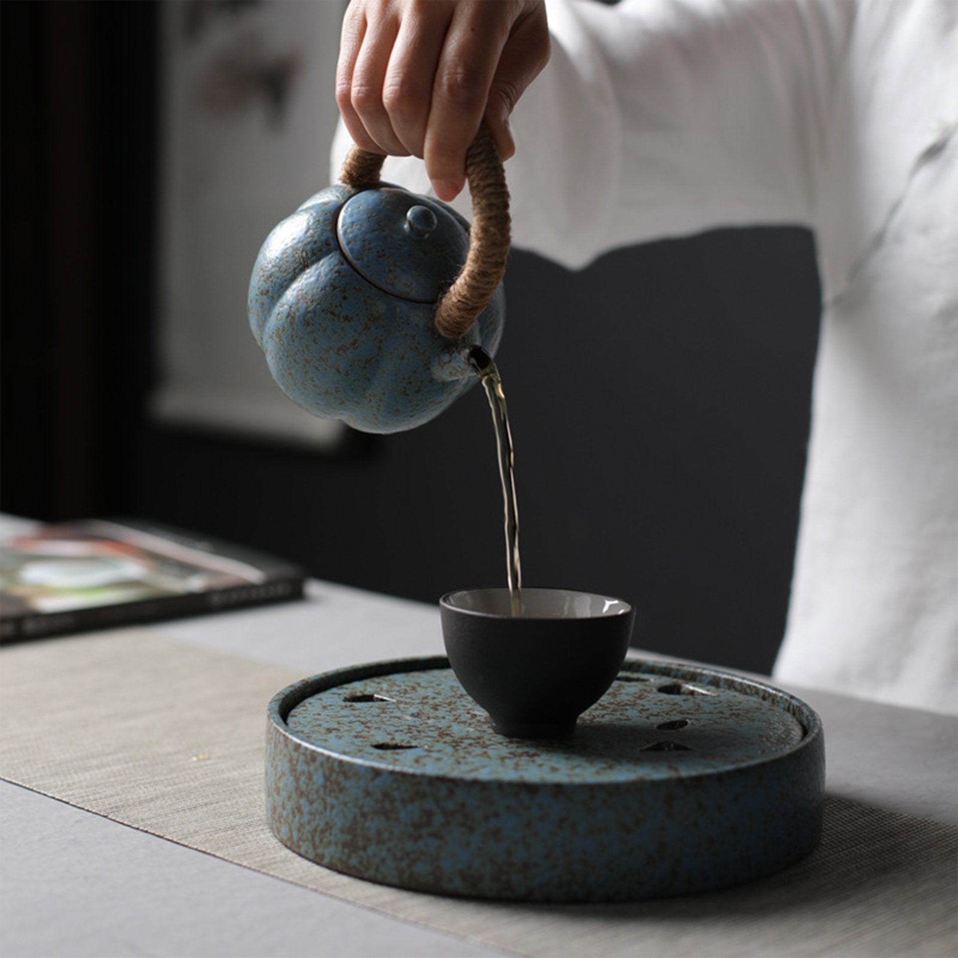 Hand pouring tea from a blue teapot into a black cup on a speckled blue tray.
