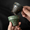 Two hands holding green and dark textured teacups close together, gold flecks visible.