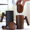 Black and brown cylindrical tea mugs with wooden handles and perforated lids on a fabric mat.Person lifting a brown tea filter from a black mug, showcasing dripping tea, with a wooden handle visible.