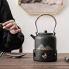 Person in black pouring tea from a black kettle on a stove into a cup