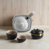 A matte black teapot with a wooden detail, two matching cups, and a light gray carrying case.