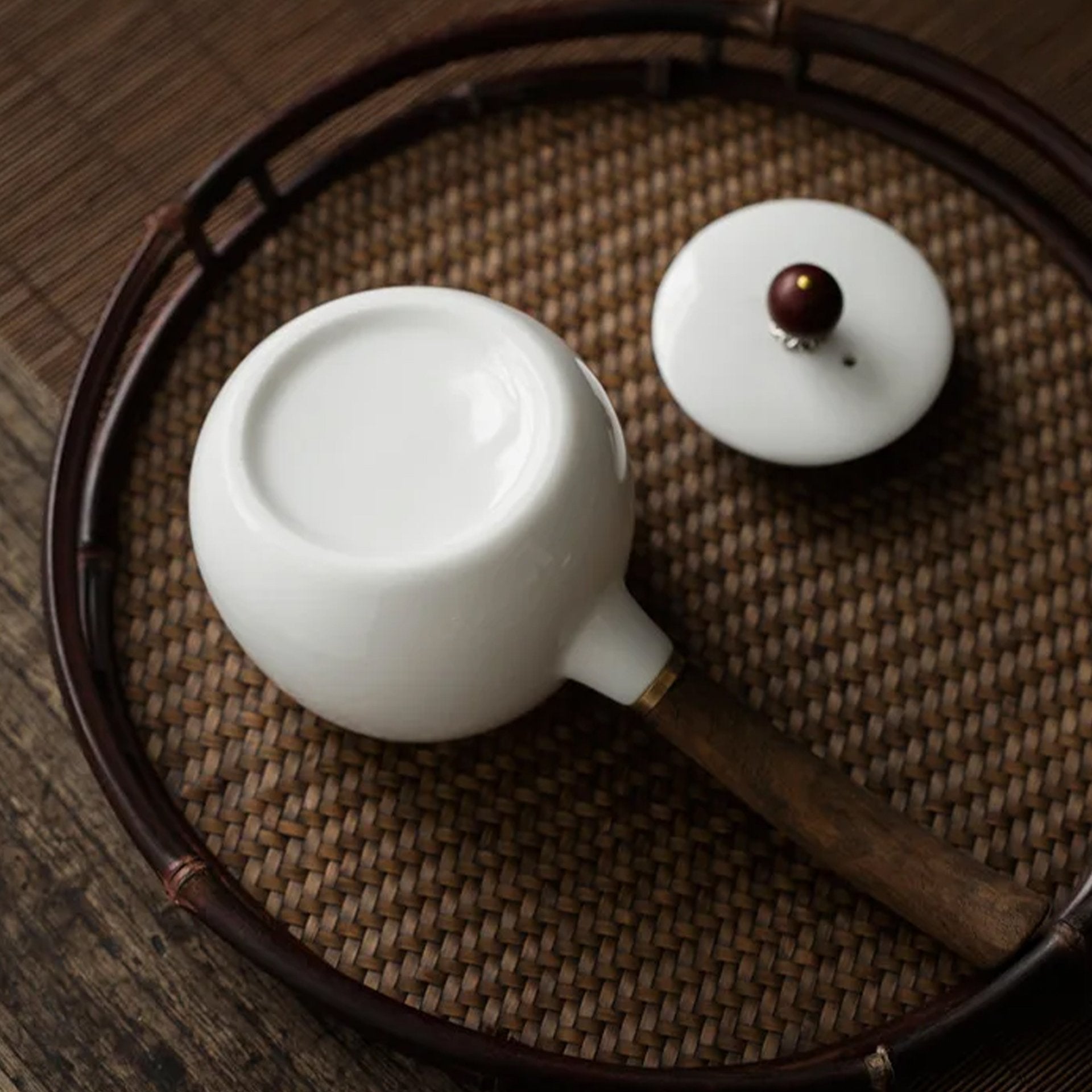 Overhead view of a white teapot with wooden handle on a woven tray with its lid off.